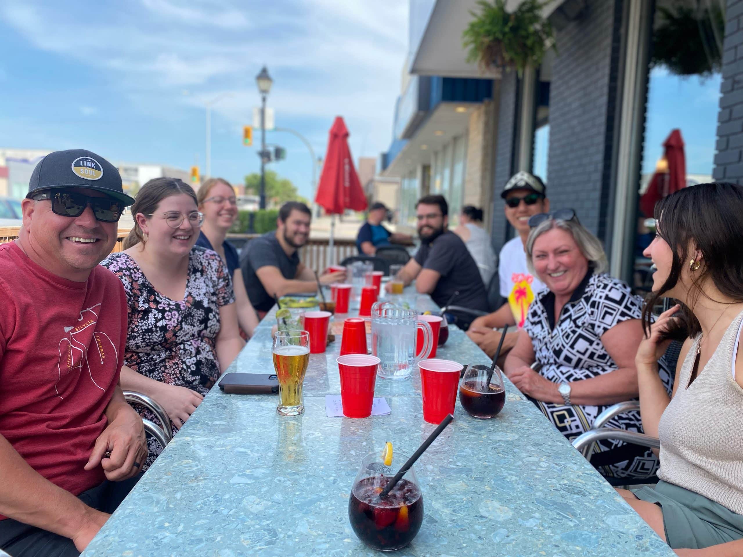 Group of happy people in summer clothing and sunglasses sitting on an outdoor patio having drinks.
